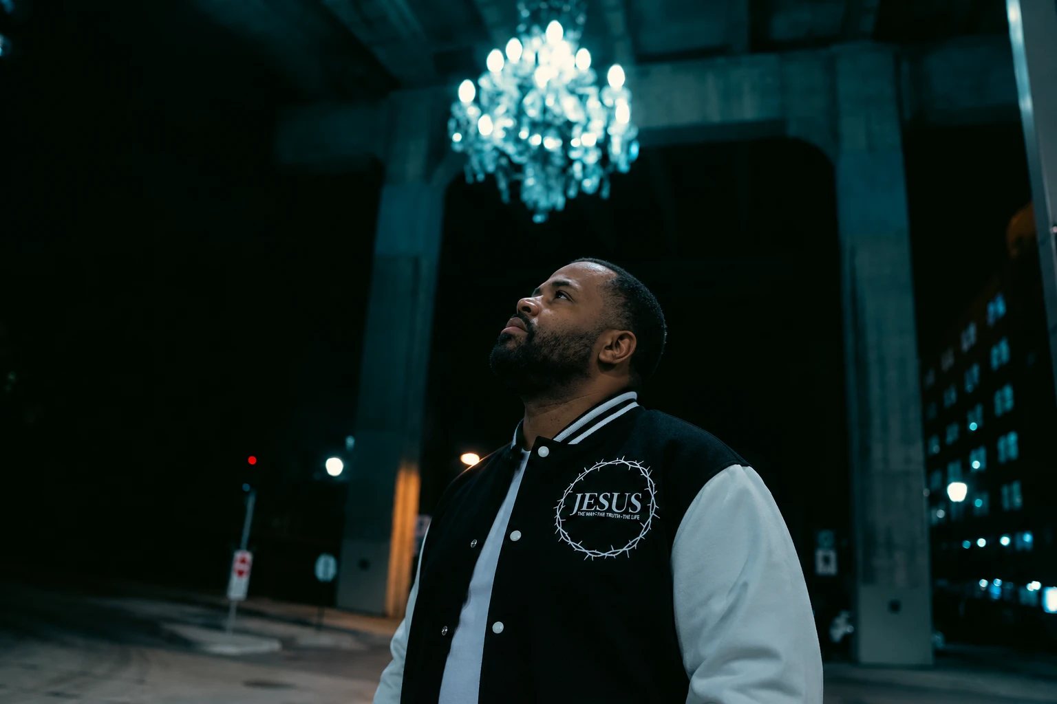 A man in the black Jesus varsity jacket looking up at a glowing chandelier